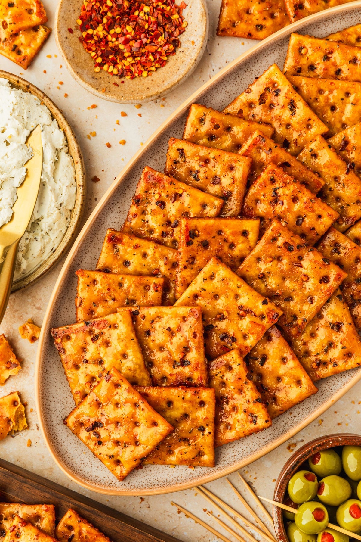 A beige platter of hot honey seasoned saltines next to brown bowls of boursin, red pepper flakes, and green olives on a tan counter.