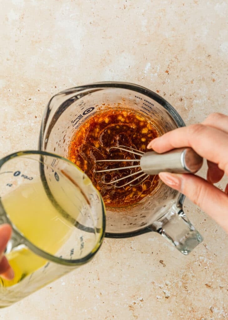 A hand pouring a measuring cup of oil into a measuring cup of dressing.