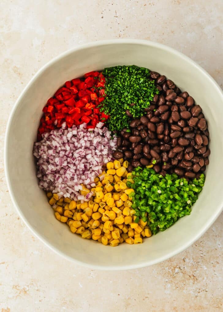 A white bowl of corn, beans, onions, peppers, jalapenos, and chives on a stone counter.