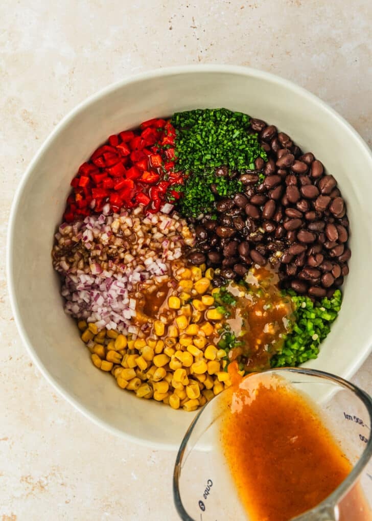 A hand pouring a measuring cup of dressing into a white bowl of veggies, corn, and beans.