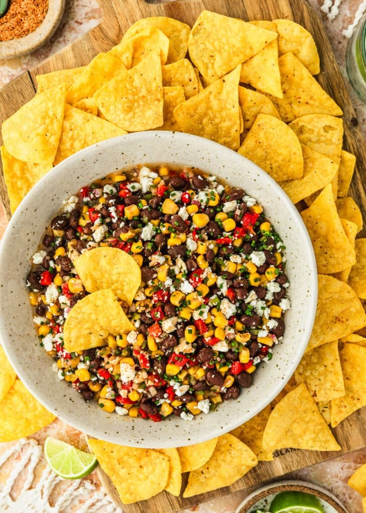 A white bowl of black bean and corn dip on a wood board of chips next to a white bowl of limes and brown bowl of Tajin on an orange marble table.