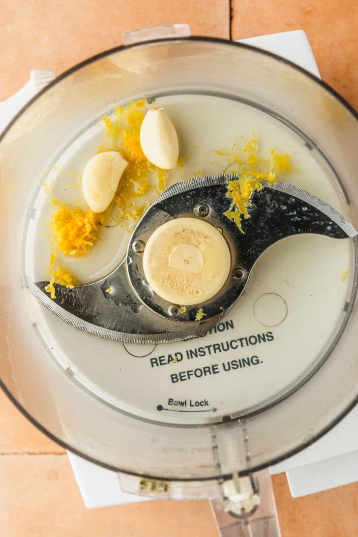 A food processor with garlic cloves and lemon zest on an orange counter.