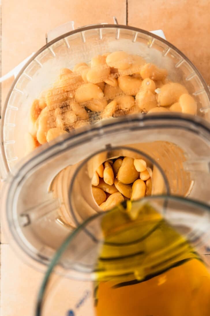 A measuring cup drizzling oil into a food processor of beans.