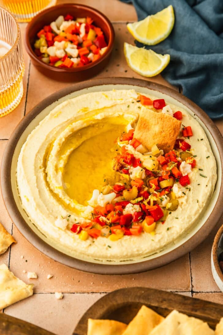 A beige platter of butter bean dip with giardiniera on top next to a blue linen, glasses of beer, and wood tray of crackers on an orange tile counter.