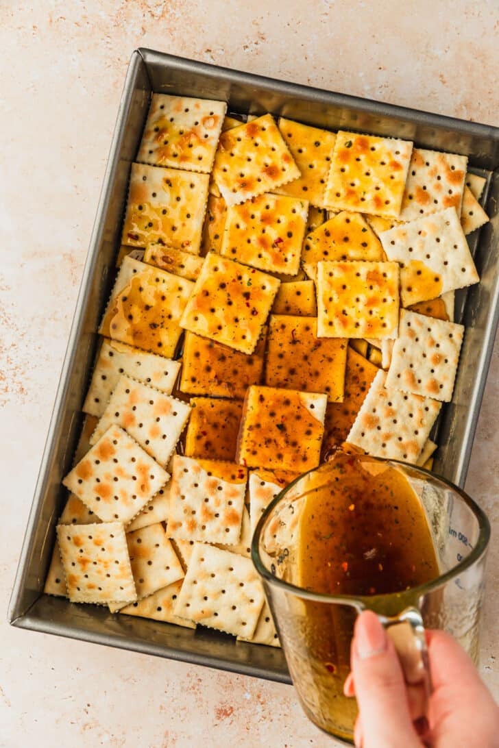 A hand pouring a measuring cup of glaze over a pan of crackers.