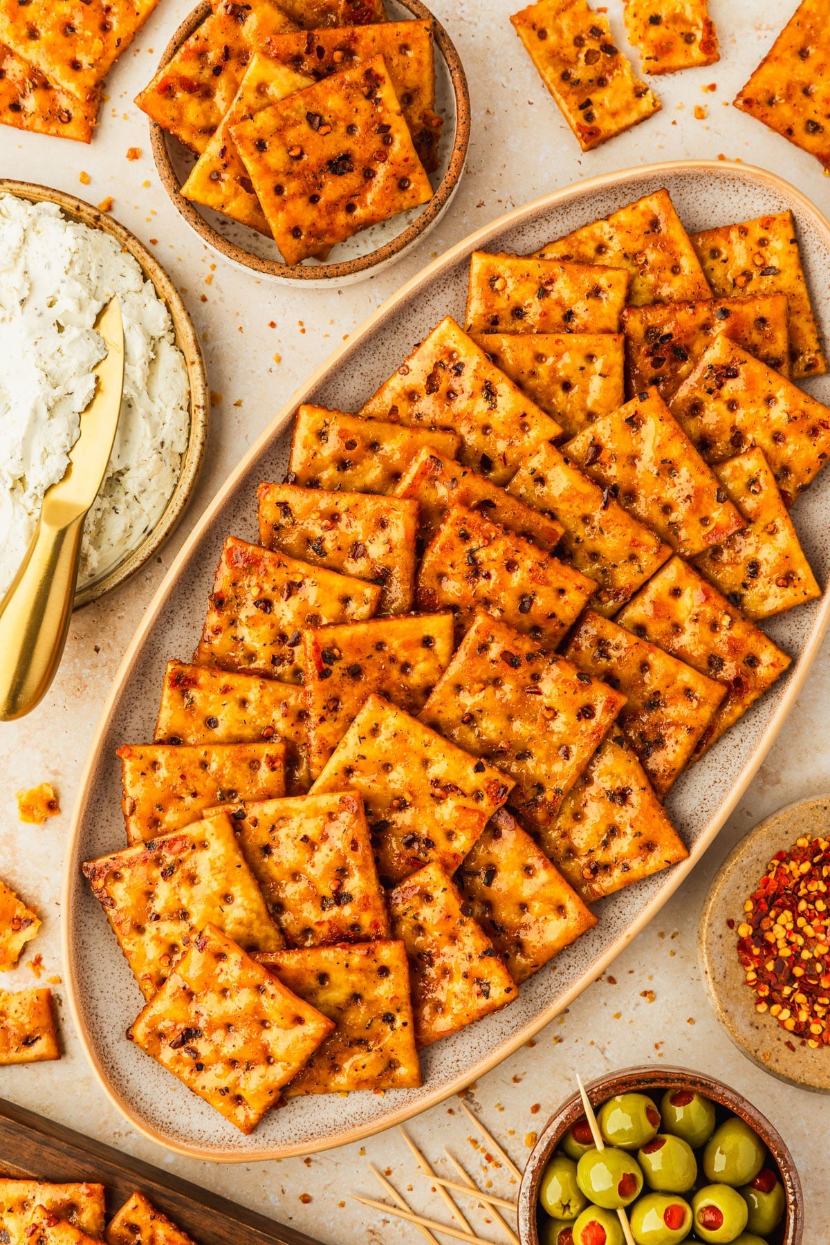 A beige platter of hot honey seasoned saltines on a tan counter next to brown bowls of crackers, boursin, green olives, and red pepper flakes.
