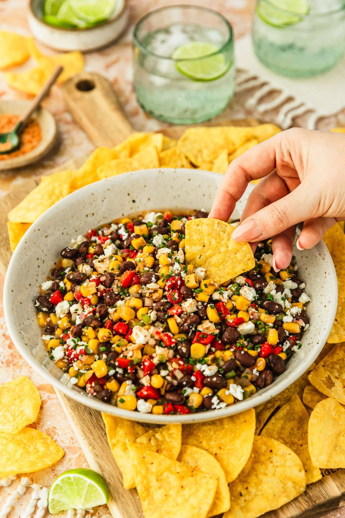 A hand dipping a corn chip into a white bowl of black bean and corn dip with feta on a wood board of chips next to glasses of ranch water, a white bowl of limes, and a brown bowl of Tajin on an orange marble counter.