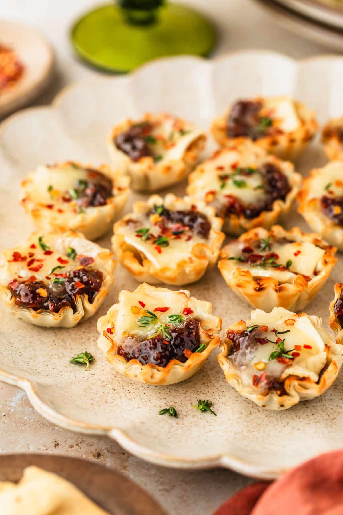 A beige platter of bacon jam and brie phyllo cups next to an orange linen, brown bowl of pepper flakes, and green bowl on a tan marble table.