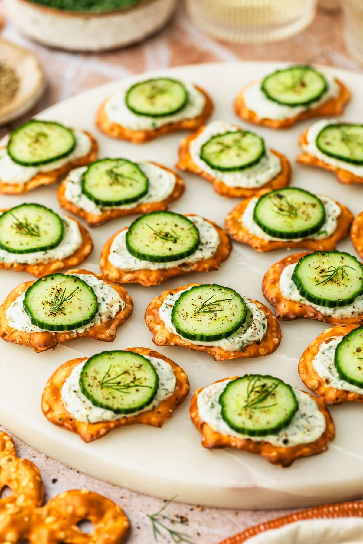 A white platter of cucumber cream cheese bites on pretzels next to an orange striped linen, glasses of white wine, and a brown bowl of pepper on an orange marble counter.