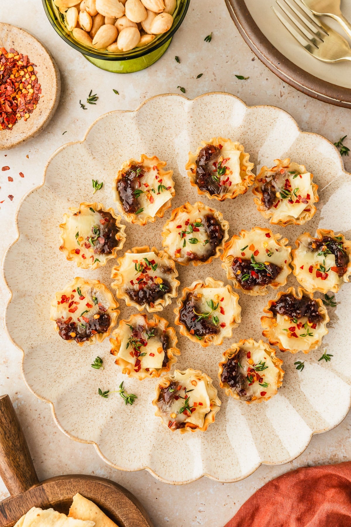 A beige platter of bacon jam and brie phyllo cups next to an orange linen, green bowl of almonds, brown bowl of red pepper flakes, and a wood board of crackers on a tan marble counter.