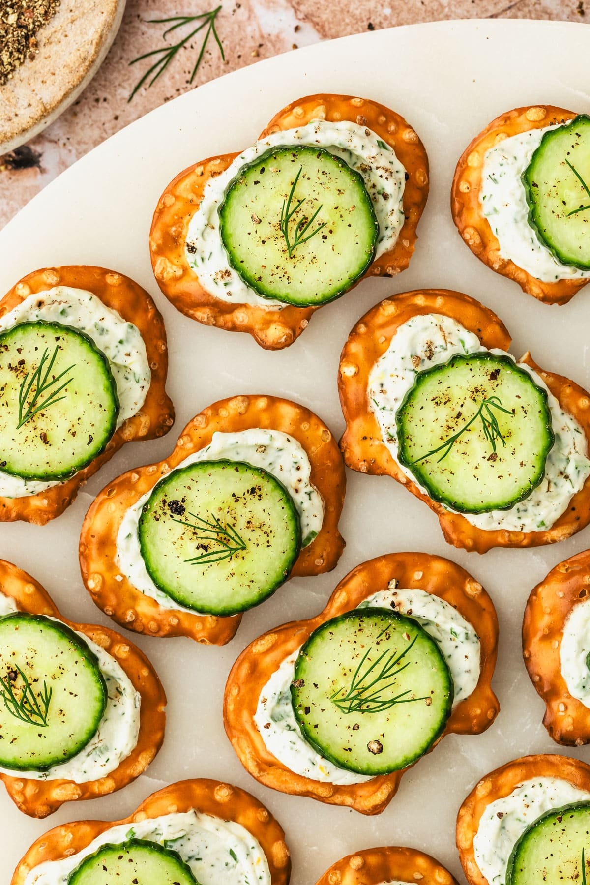 A white platter of cucumber cream cheese bites on pretzels next to a brown bowl of pepper on a dark orange-brown table.