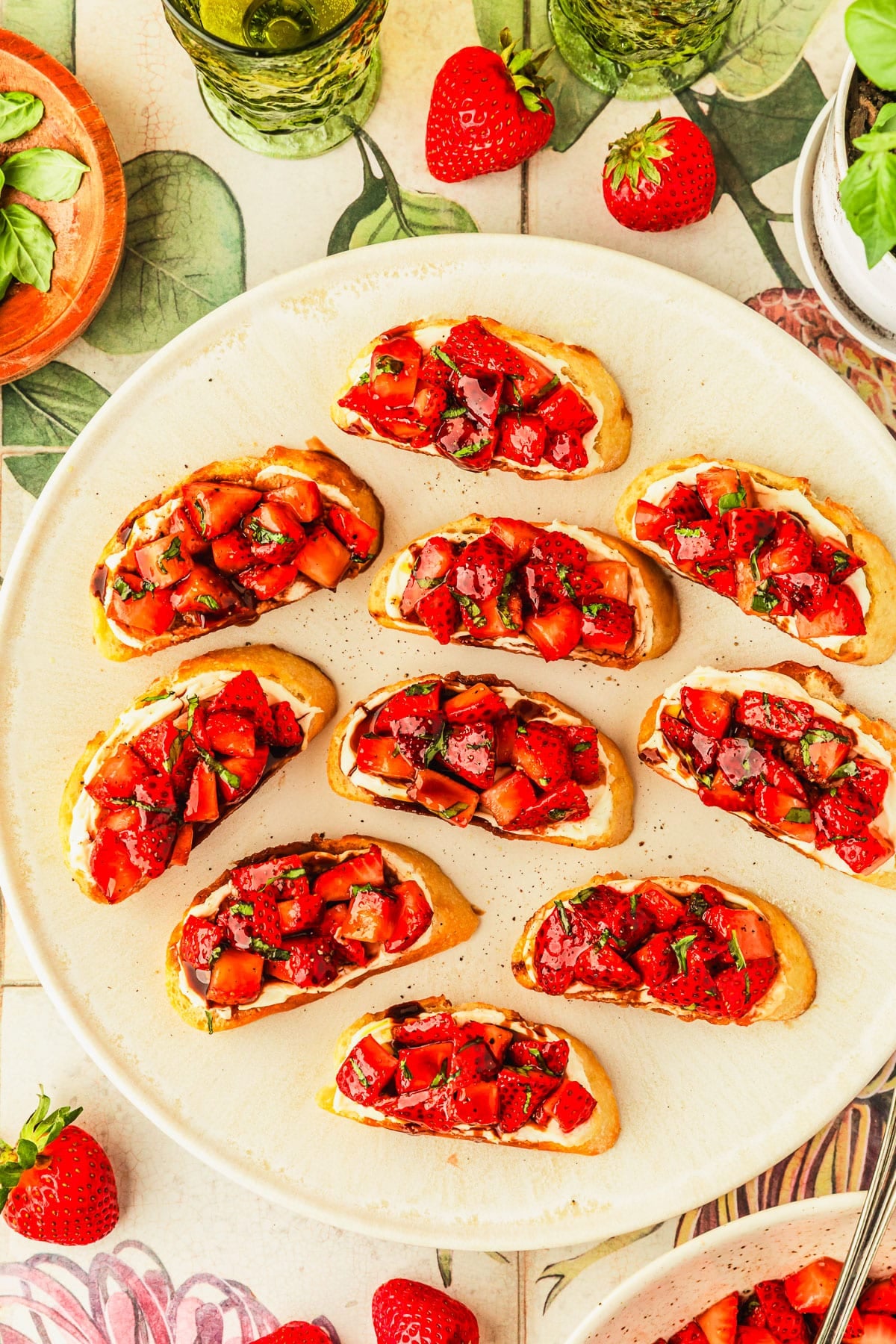 A white platter of strawberry bruschetta next to a wood bowl of basil, strawberries, and green glasses of white wine on a green, pink, and white floral counter.