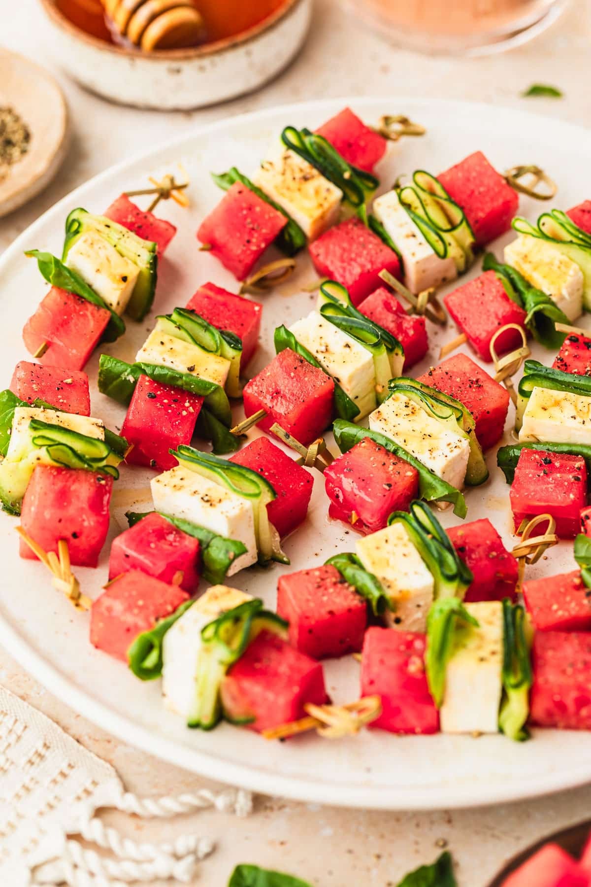 A white platter of watermelon feta skewers with hot honey next to glasses of rose, a white napkin, and a white bowl of hot honey on a tan counter.