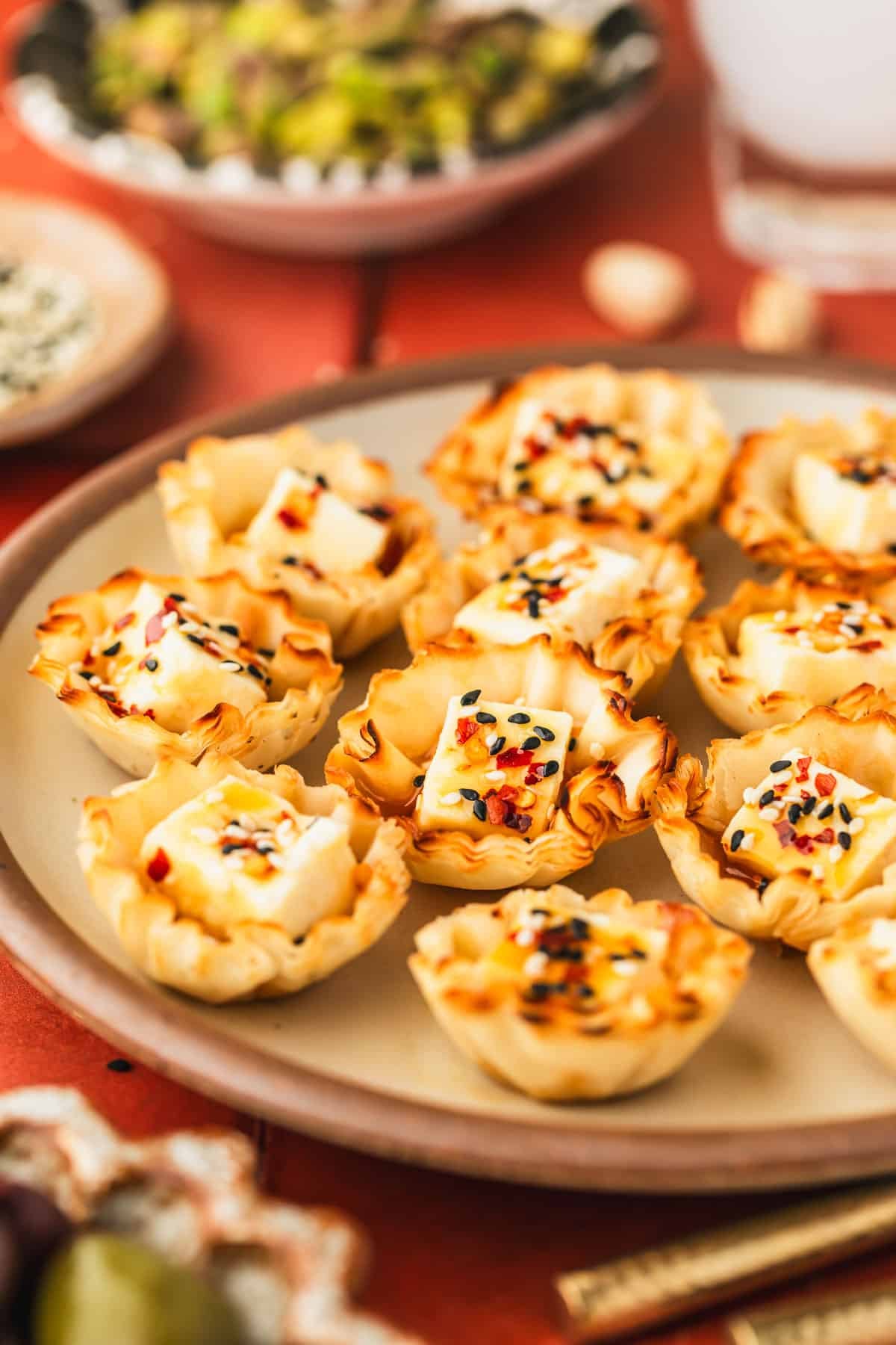 A beige platter of feta phyllo cups next to a glass of ouzo, a brown bowl of sesame seeds, and a black bowl of pistachios on a red tiled counter.