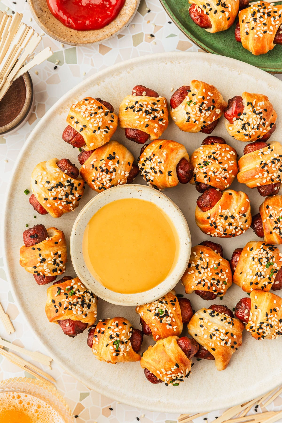 A white platter of air fryer pigs in a blanket on a white terrazzo counter next to a green plate of pigs in a blanket, glass of beer, brown bowl of toothpicks, and brown bowl of ketchup.