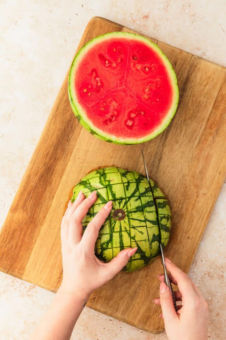 A hand using a knife to slice a watermelon into cubes.