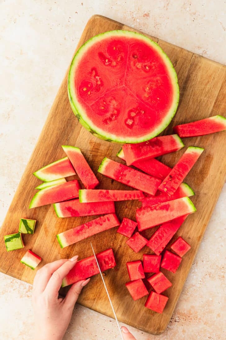 Hands using a knife to slice watermelon sticks into cubes on a wood board.