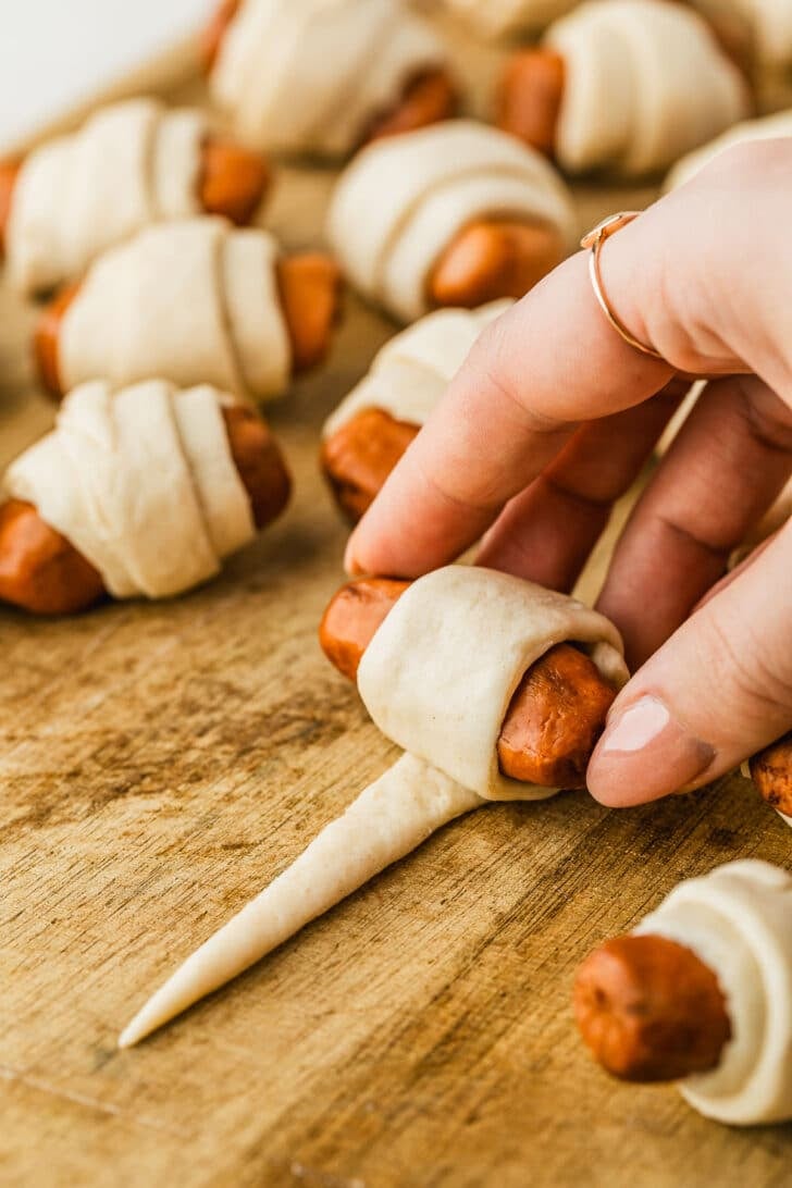 A hand rolling a mini sausage in dough on a wood board.