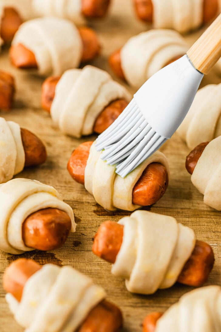 A pastry brush brushing egg wash on raw pigs in a blanket.