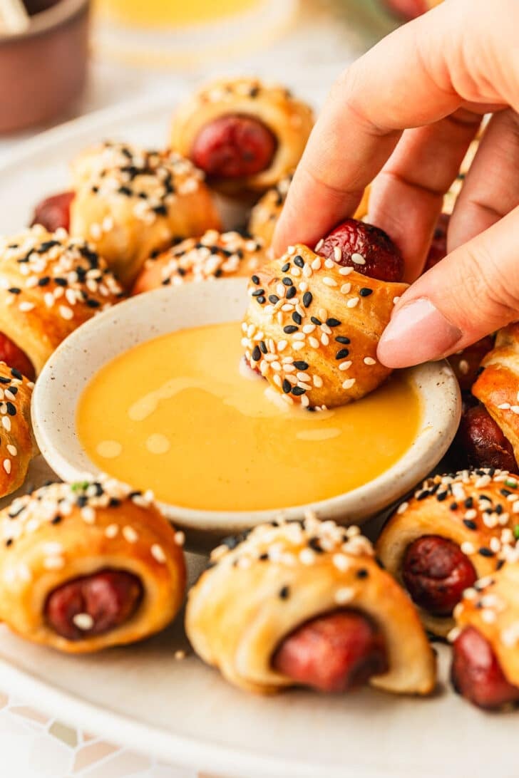 A hand dipping a pig in a blanket in mustard on a white platter of air fryer pigs in a blanket.