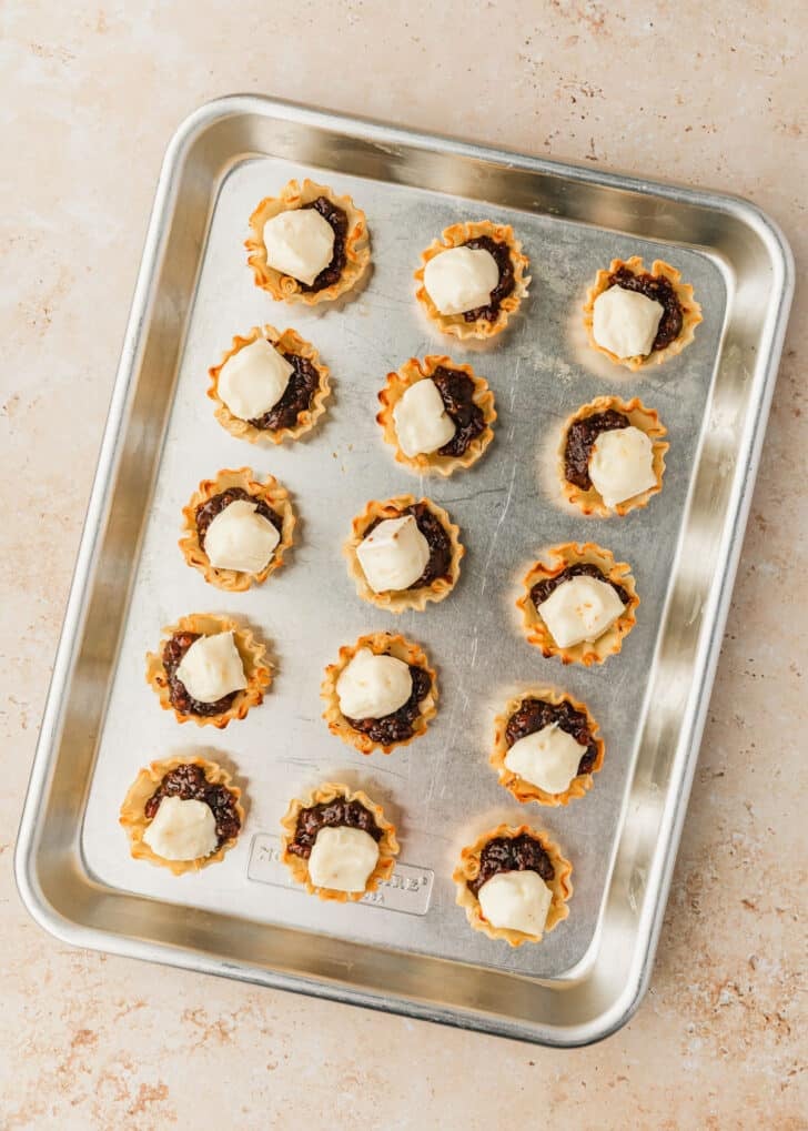A pan of filo shells with jam and cheese on a tan counter.