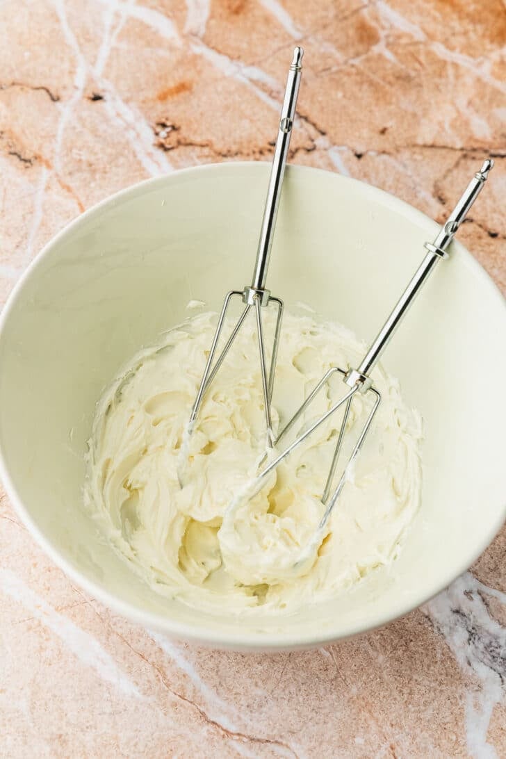 A white bowl of whipped cream cheese with beaters on an orange marble counter.
