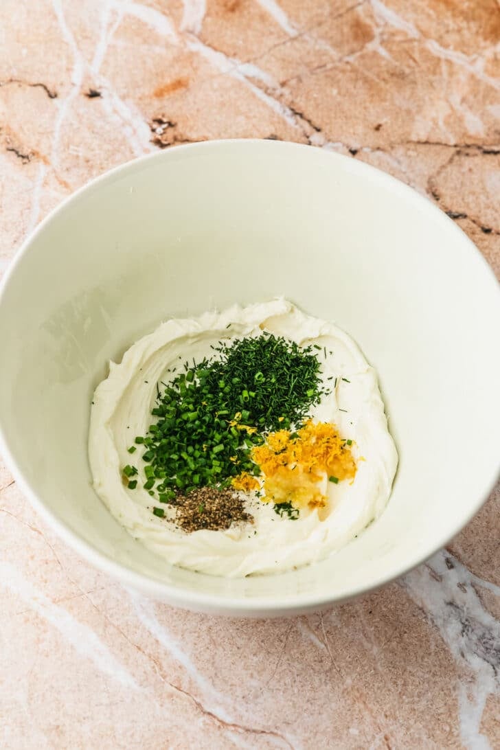 A white bowl of cream cheese, herbs, garlic, and lemon on an orange counter.