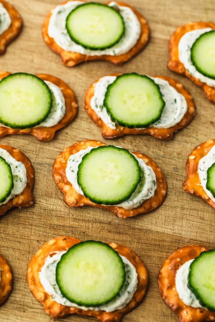 A wood board of cucumber cream cheese bites on pretzel crisps.