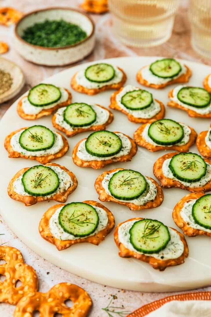 A white platter of cucumber cream cheese bites on pretzels next to glasses of white wine, a white bowl of chives, and a brown bowl of pepper on an orange stone background.