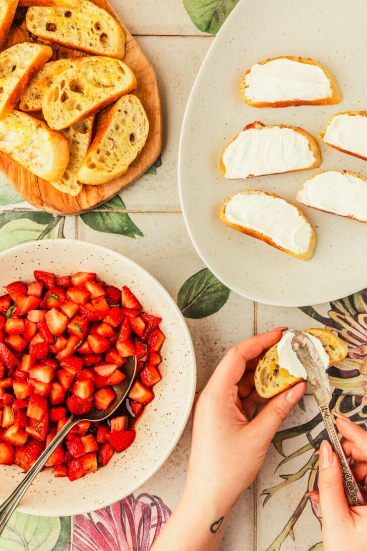 A hand using a knife to spread mascarpone on crostini next to a wood board of crostini, white platter of crostini with cheese, and a white bowl of berries.