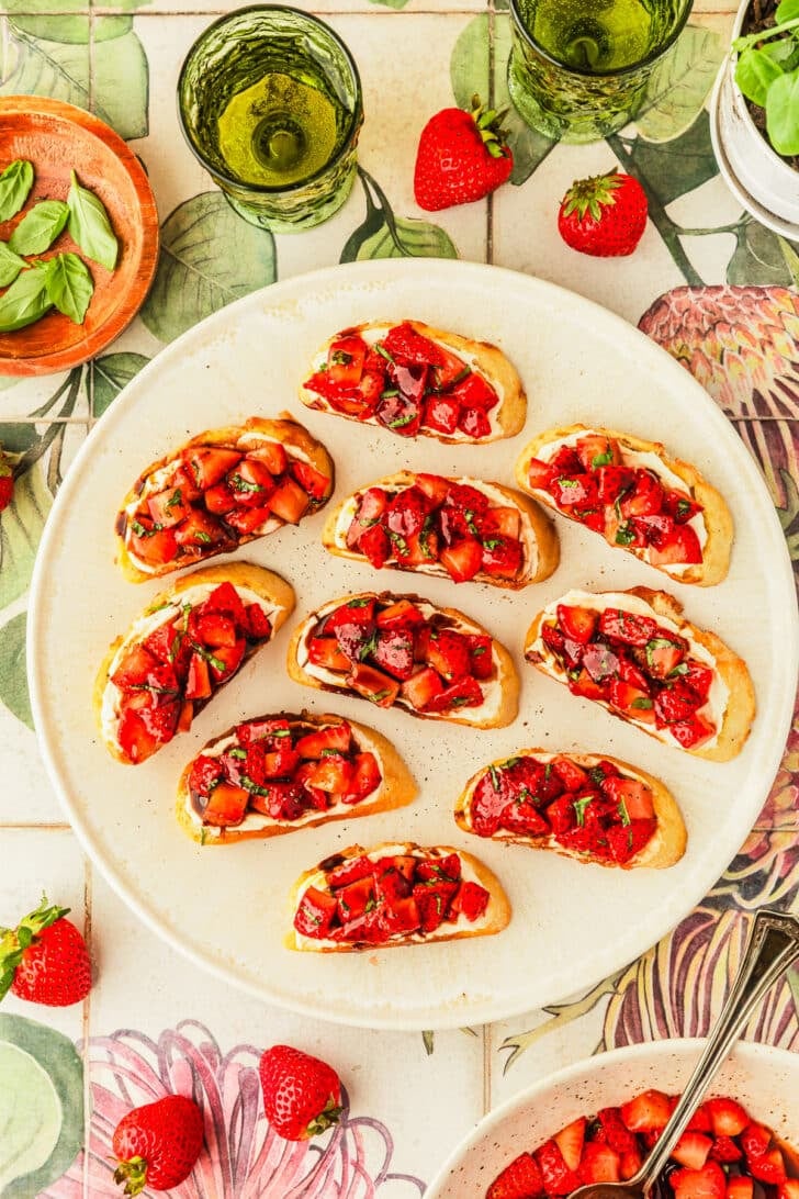 A white platter of strawberry balsamic bruschetta with mascarpone next to a white bowl of berries, strawberries, and green glasses of white wine on a green and white floral counter.