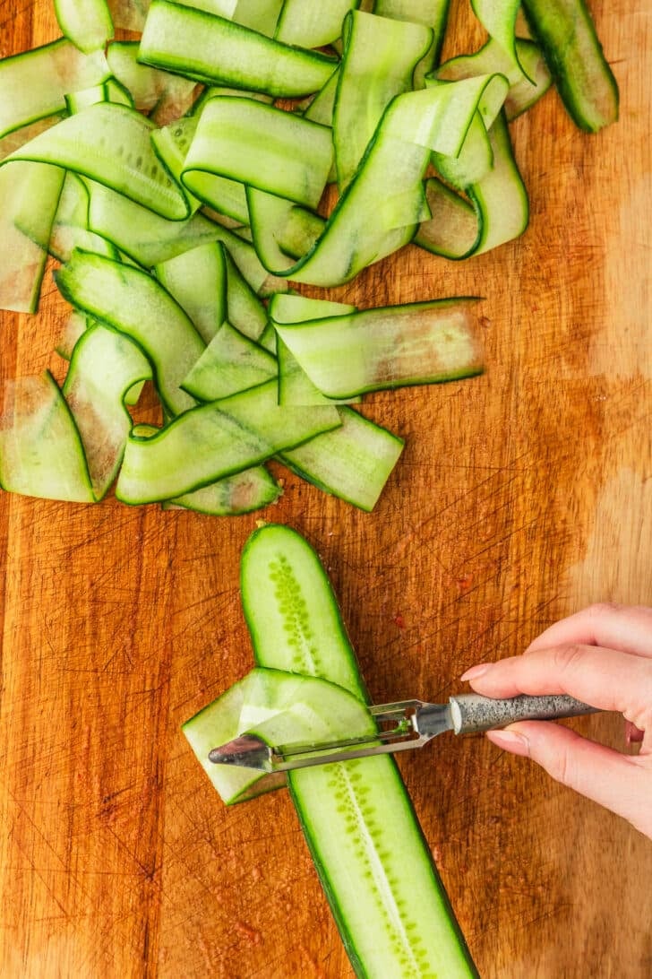 A hand using a vegetable peeler to make cucumber ribbons on a wood board.