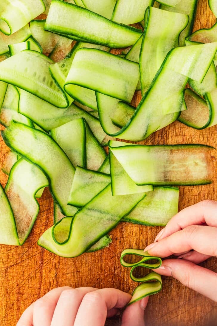 Hands assembling cucumber ribbons on a wood board.