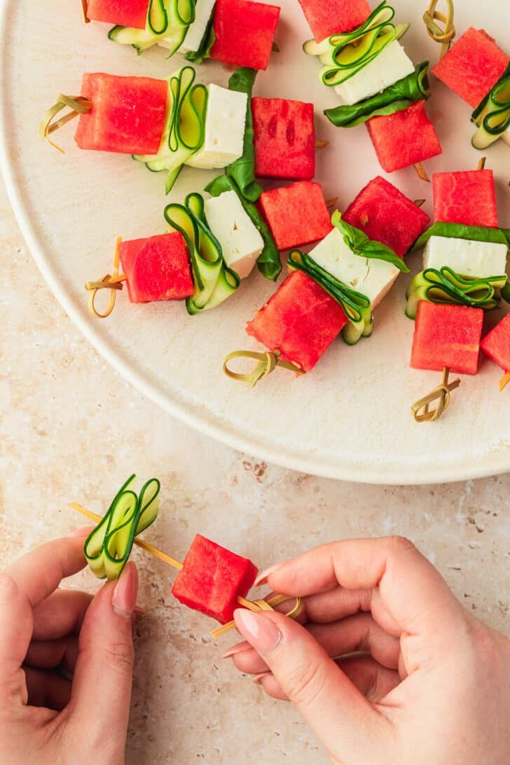 Hands adding a cucumber ribbon to a skewer next to a white plate of watermelon feta skewers.