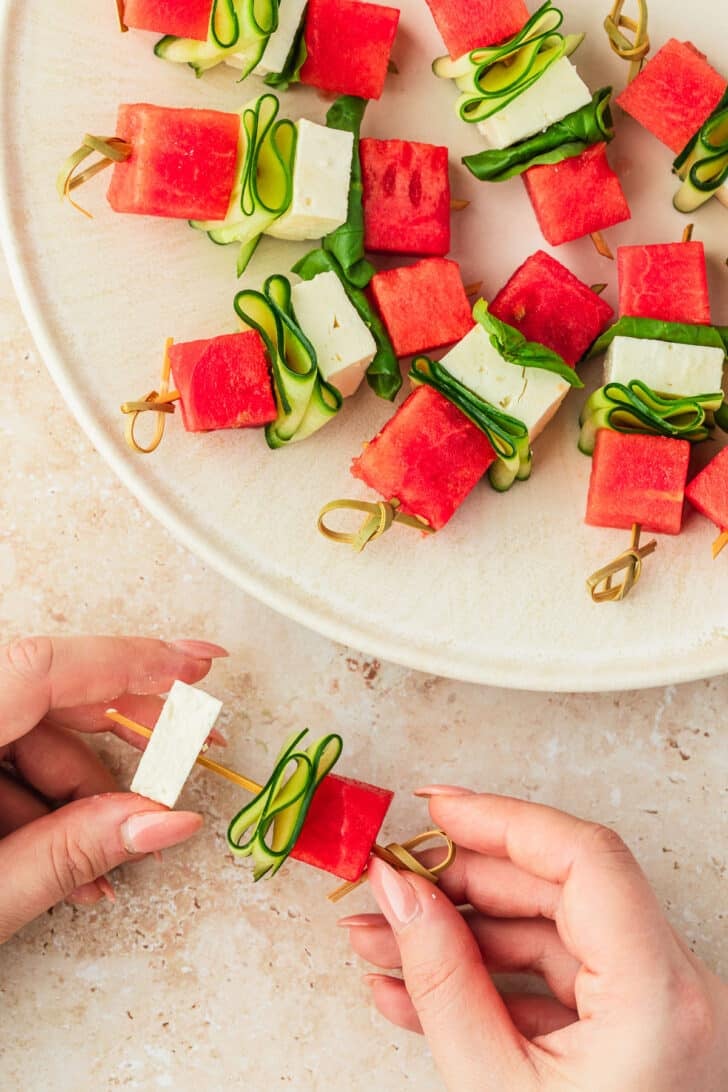 Hands adding a cube of feta to a skewer next to a white plate of watermelon feta skewers.