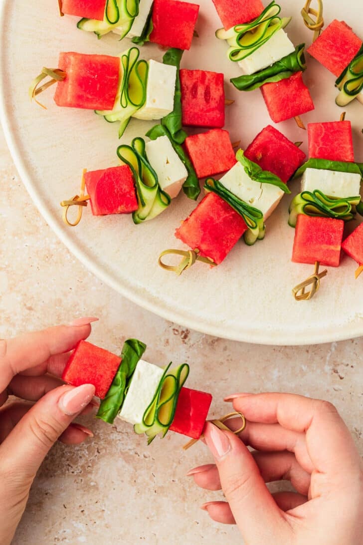 Hands assembling watermelon feta skewers on a tan counter.