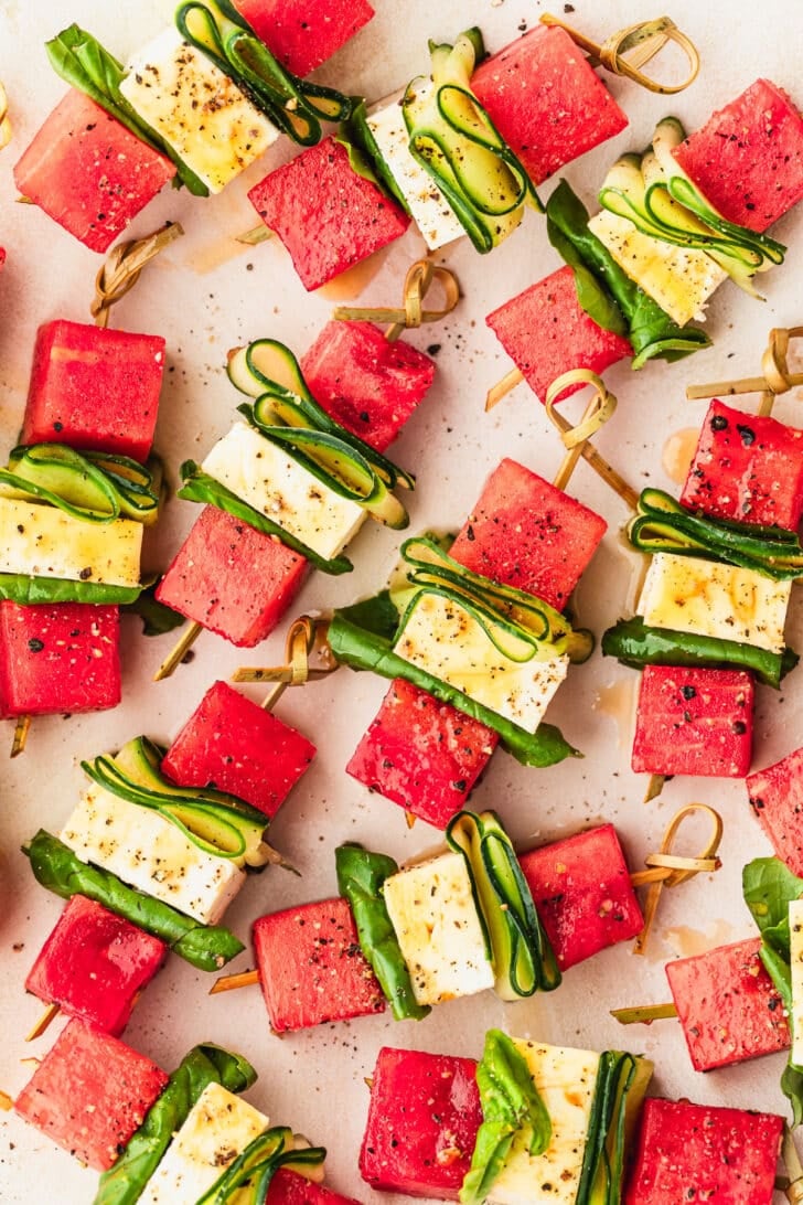 A white platter of feta watermelon skewers with cucumbers, basil, and hot honey.