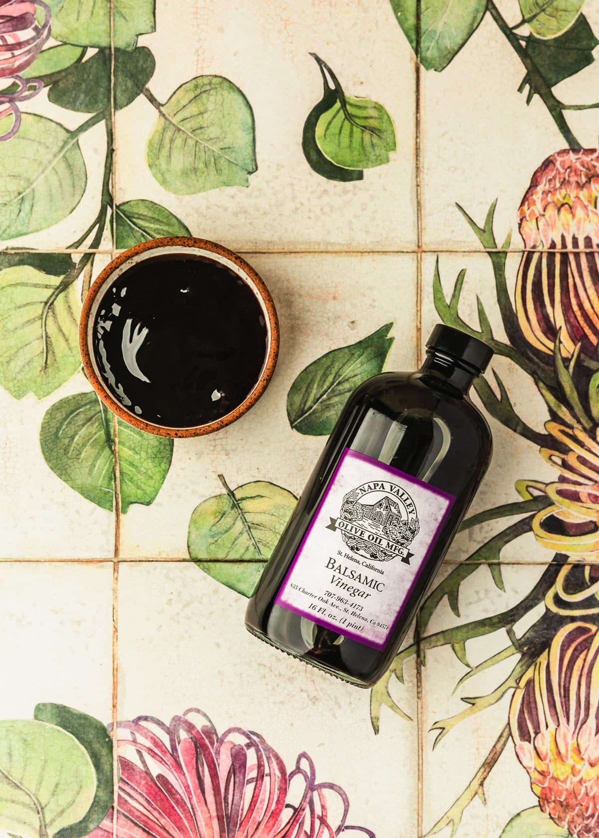 A white bowl of balsamic glaze and a bottle of balsamic vinegar on a pink, white, and green floral counter.