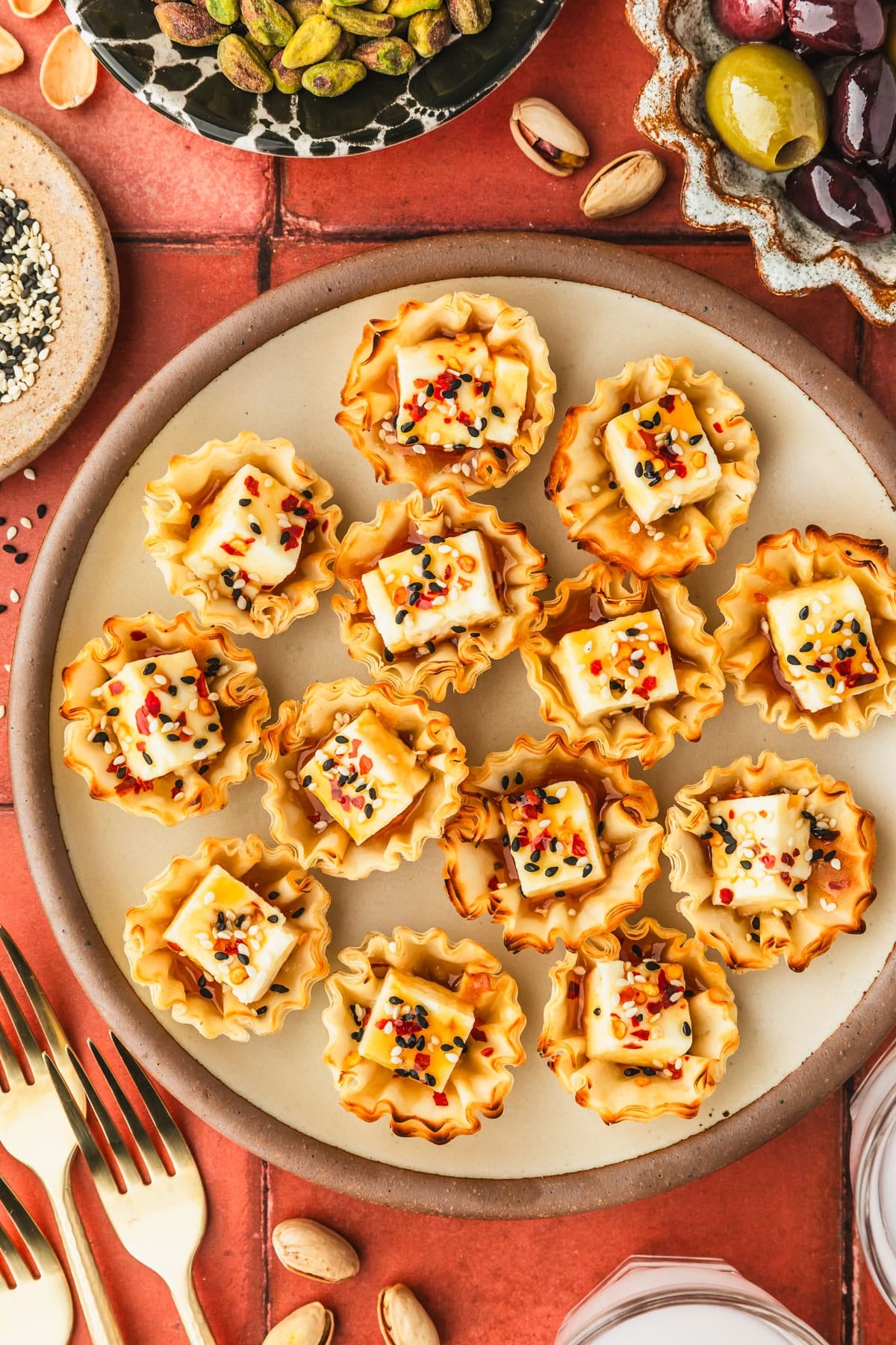 A stoneware plate of feta phyllo cups with hot honey next to brown bowls of olives and sesame seeds, glasses of ouzo, and a black bowl of pistachios on a red tiled counter.