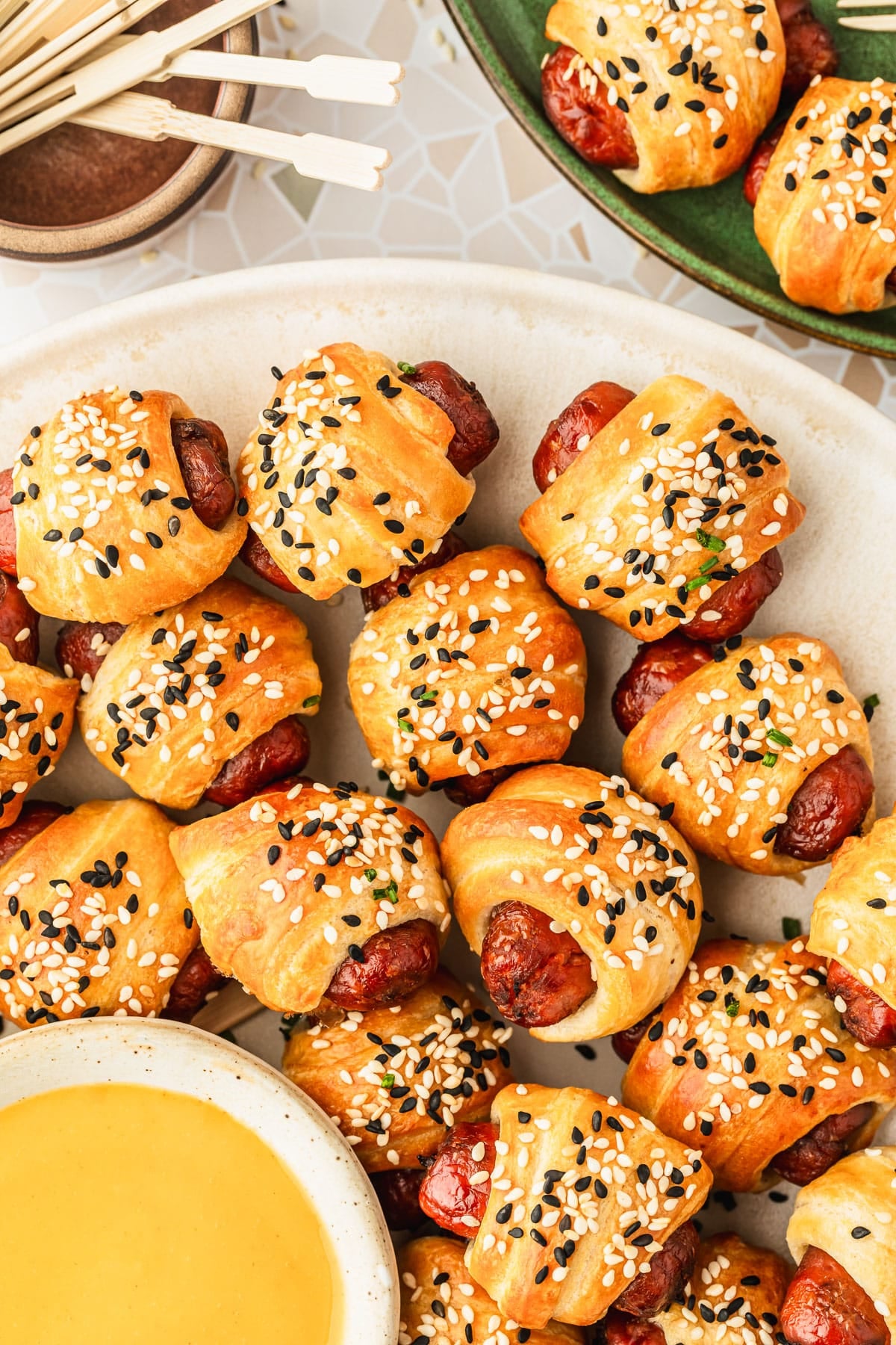 A white platter of air fryer pigs in a blanket and a white bowl of honey mustard on a beige terrazzo counter next to a brown bowl of toothpicks and green plate.