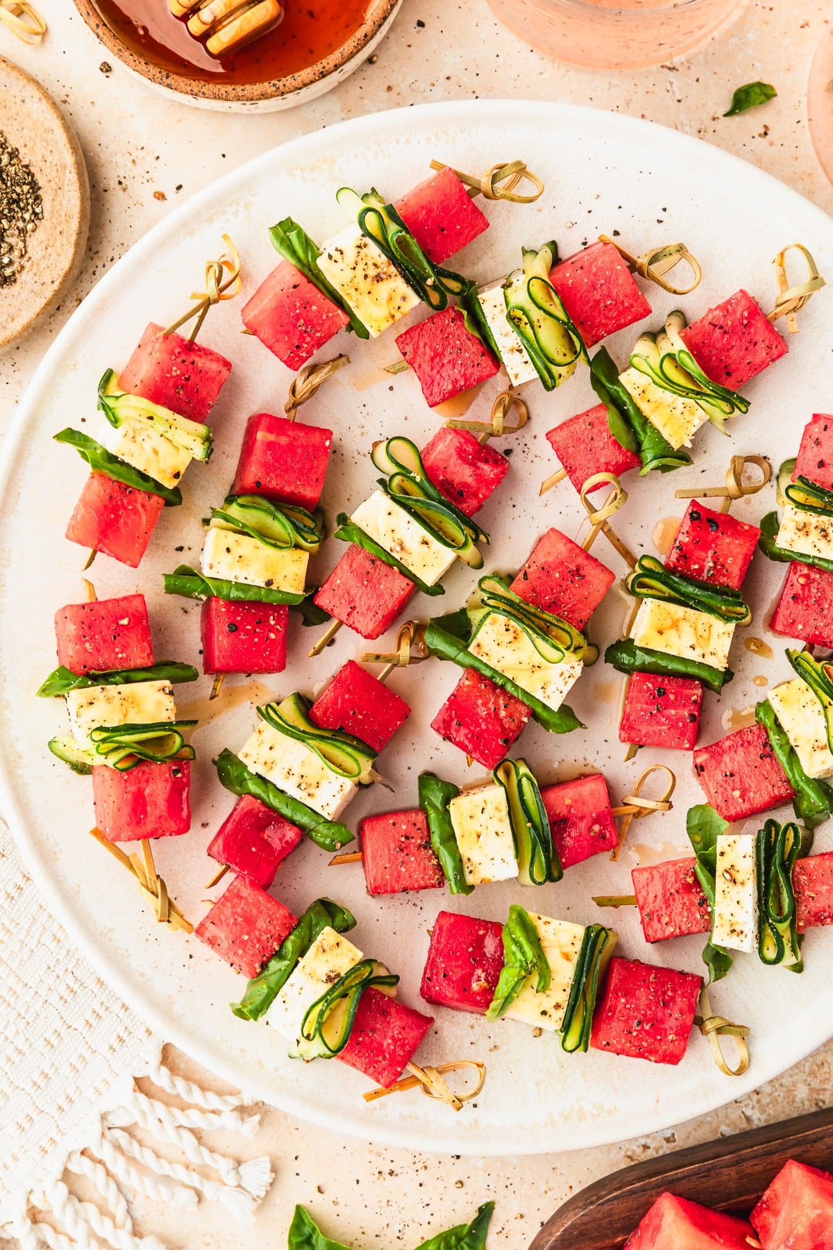 A white platter of feta watermelon skewers with cucumber, basil, and hot honey on a tan counter next to glasses of rose, a brown bowl of hot honey, and a white fringe napkin.