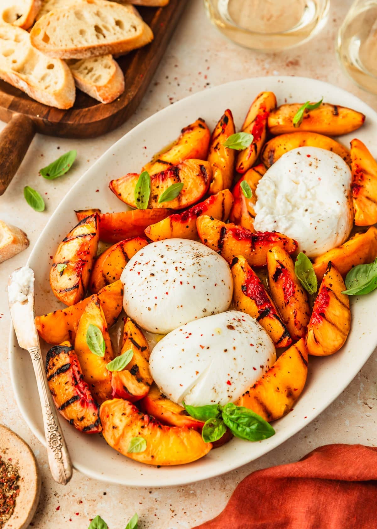 Peaches and burrata appetizer on a white platter next to glasses of wine, a wood board of bread, and an orange napkin on a tan table.