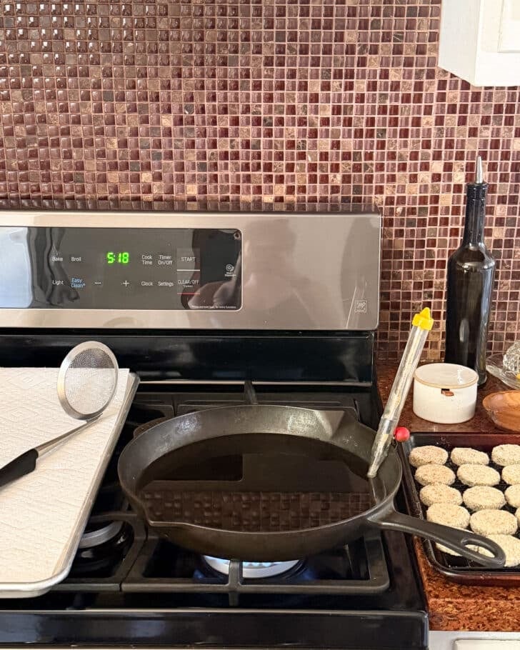 A pan of oil on a stove next to a sheet pan and a wire rack of breaded zucchini for fried zucchini.