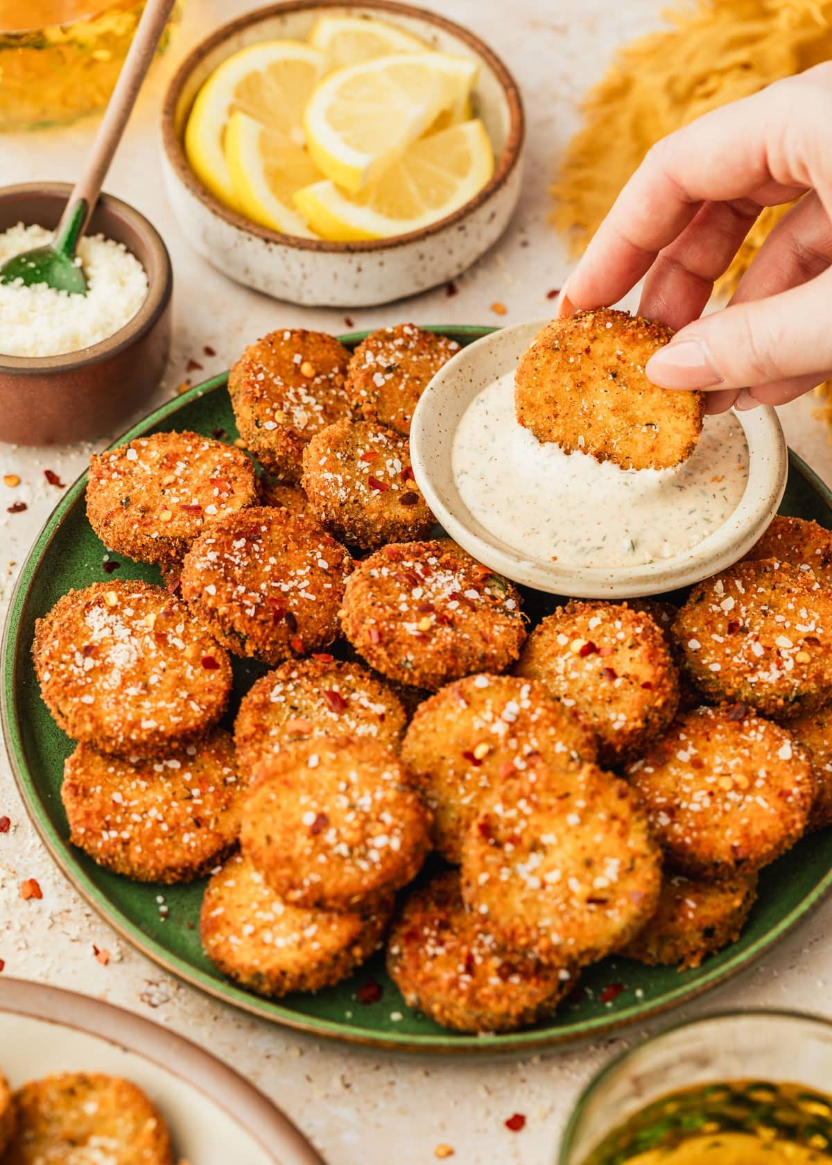 A hand dipping fried zucchini into a white bowl of ranch on a green plate of fried zucchini next to glasses of beer, a yellow linen, a white bowl of lemons, and a brown bowl of parmesan with a tan background.
