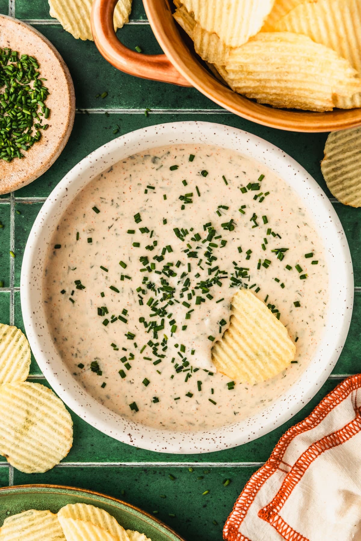 A white bowl of homemade buttermilk ranch dip on a green tiled counter next to a brown bowl of potato chips, a green plate, and a brown bowl of chives.