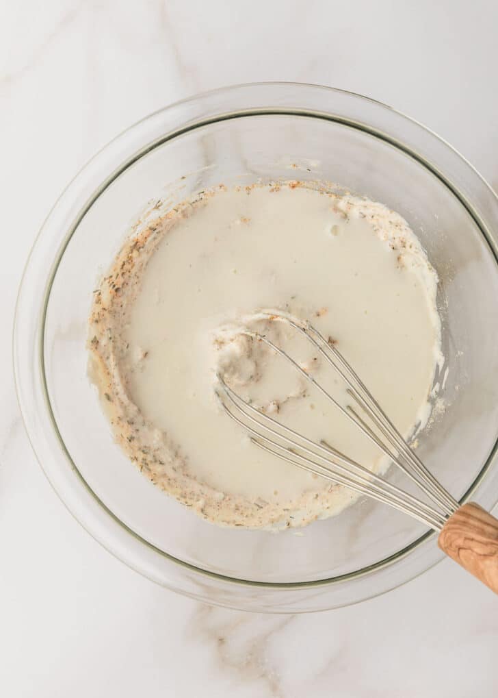 A white bowl of mayo, sour cream, seasoning, and buttermilk on a white marble table.