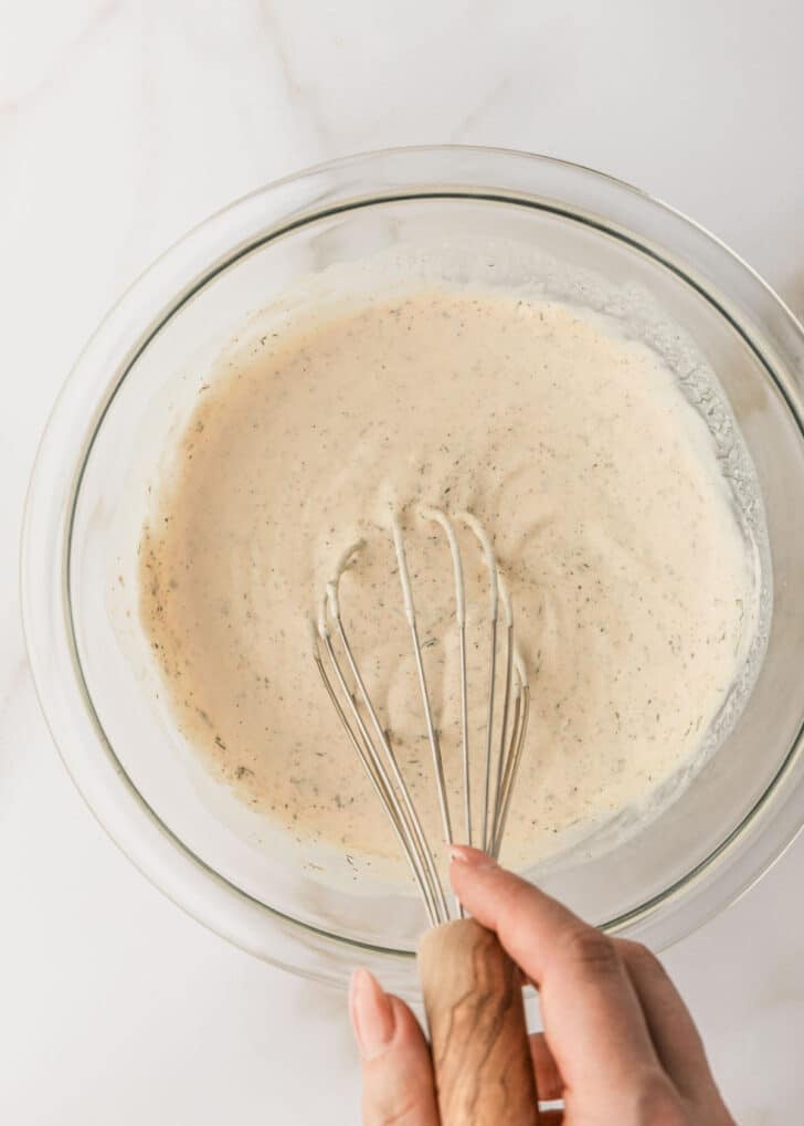 A hand using a whisk to stir homemade buttermilk ranch dip in a clear bowl.