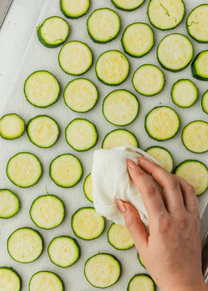 A hand using a paper towel to dry zucchini coins.