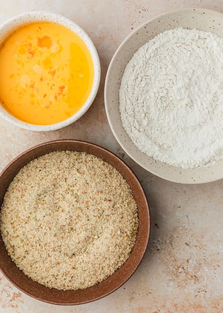 White and brown bowls of flour, breadcrumbs, and eggs on a tan counter.