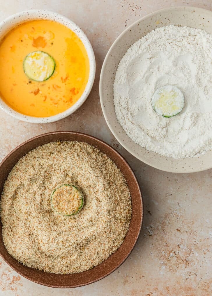 Slices of zucchini in white and brown bowls of flour, eggs, and breadcrumbs.